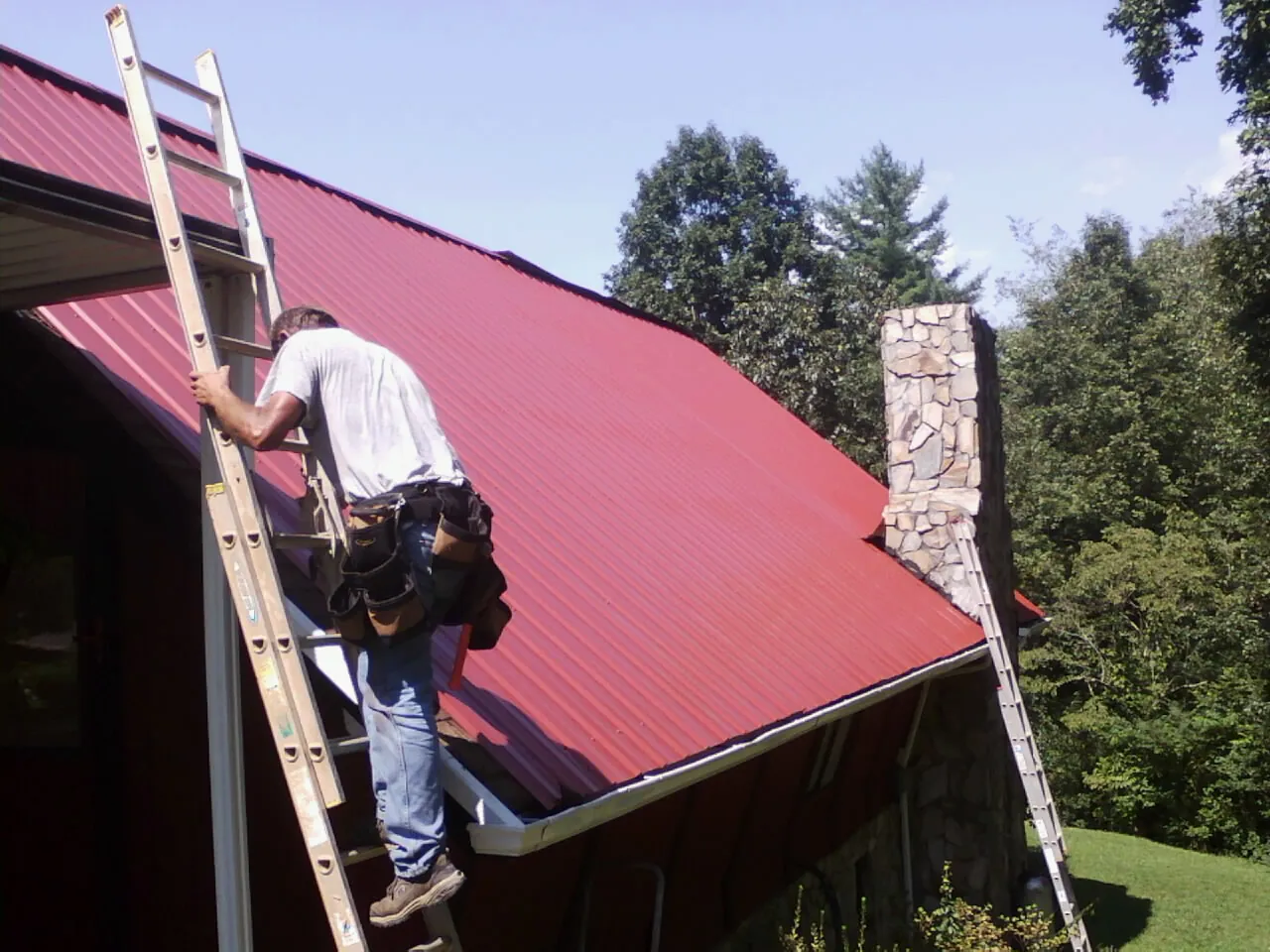 Roofer on a ladder beside a red metal roof and stone chimney — Soffit & Fascia Repair by Roofing Brothers in Stoddard